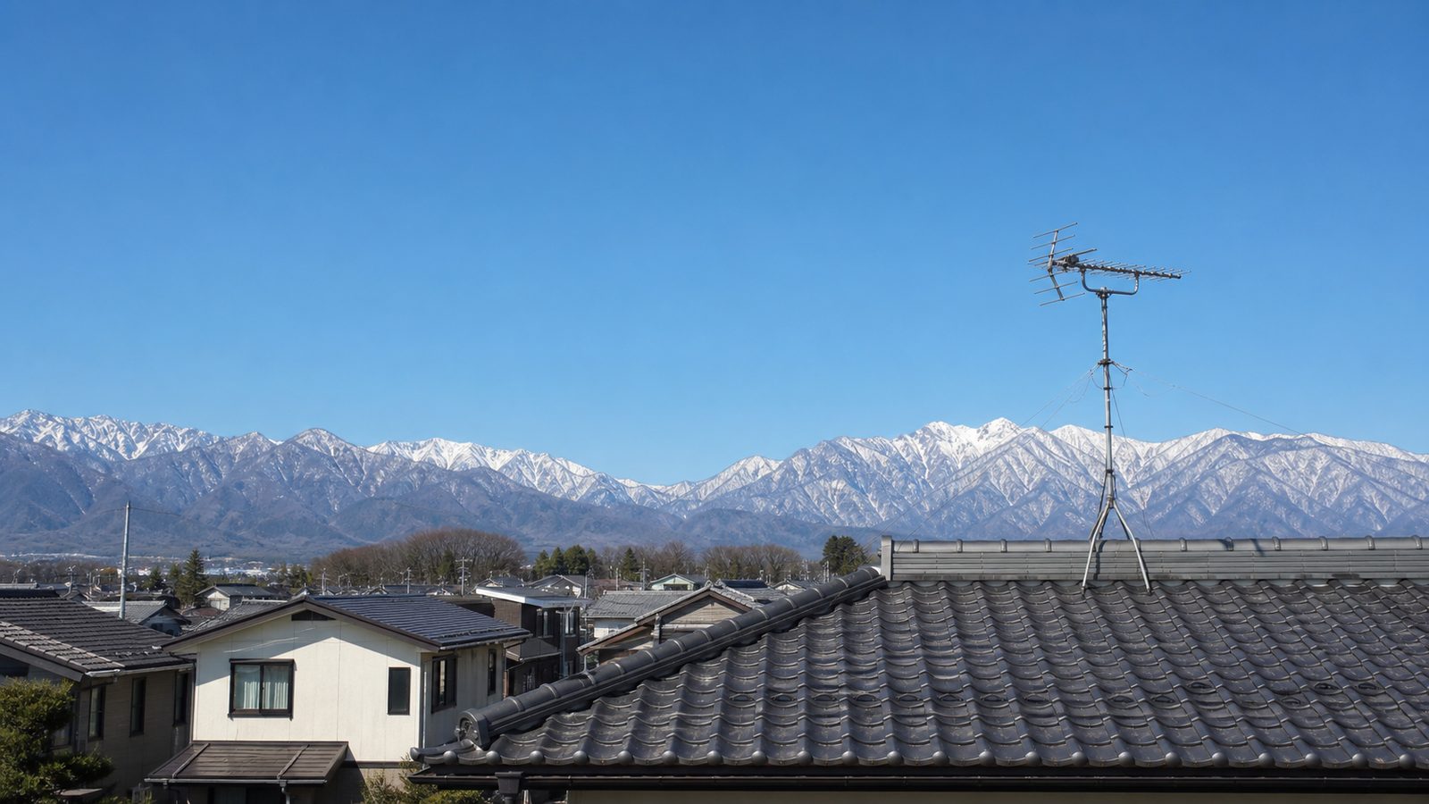 北アルプスの山並みを背景に、瓦屋根とテレビアンテナのある松本市内の住宅街